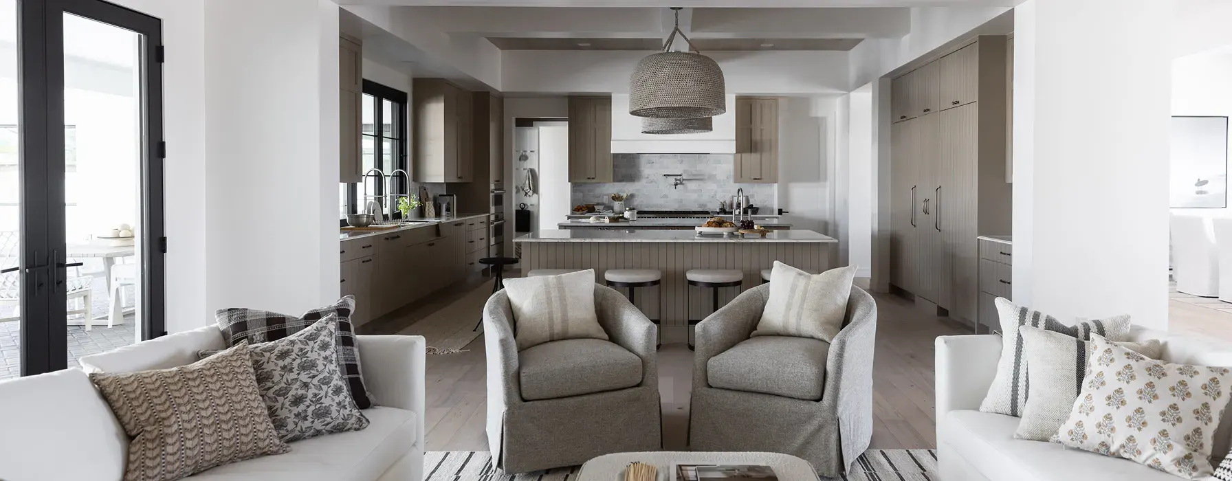 Open-plan living room and kitchen with neutral seating, two armchairs facing a coffee table, and a bright kitchen with island and bar stools in the background.